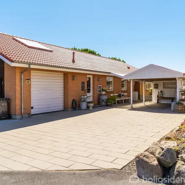 En murstensvilla med rødt tegltag har en garage og en overdækket terrasse. Haven er anlagt med sten og planter. Til venstre ses en sort garageport. Adresse: 27.