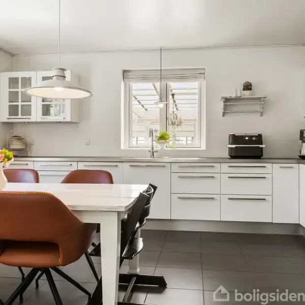 Et moderne køkken med hvide skabe fyldt med grå bordplader, spisebord med brune stole, vase med blomster og huskelampe. Vindue i baggrunden giver naturligt lys.