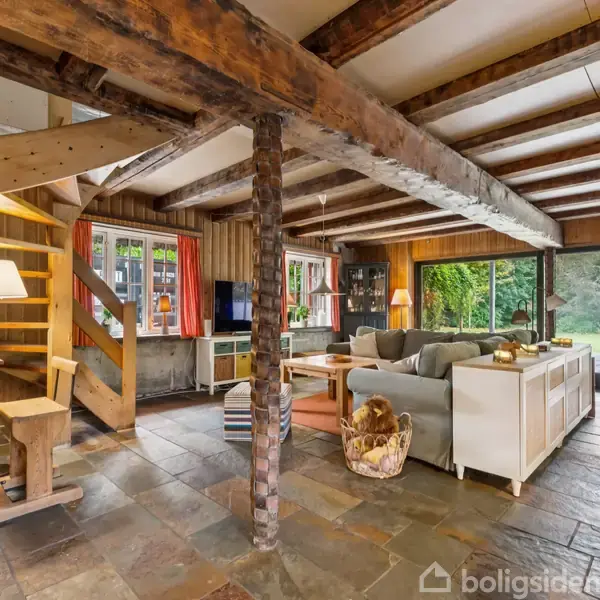 Træbjælke-stue med sofa, pejs og trætrappe. Udsigt til have gennem store glasdøre. Møbleret med tv, bogreol og lamper, hvilket skaber en hyggelig atmosfære. Flisegulv og tæppe.