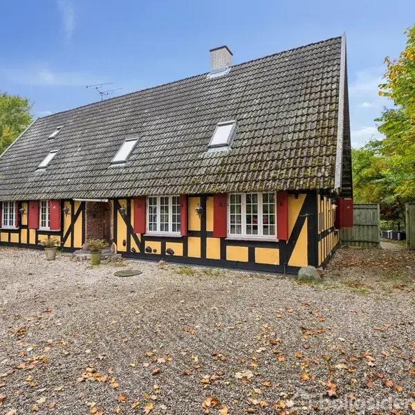 Bindingsværkshus med skæve, gule og sorte vægge står stille på en grusbelagt gårdsplads, omgivet af træer og buske i naturfyldte omgivelser under en klar blå himmel.