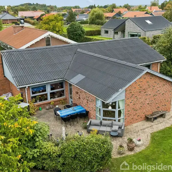 Et murstenshus med sort tag står i en grøn have med buske og træer. Der er en terrasse med møbler og en blå presenning. Omkring ligger andre huse og haver.