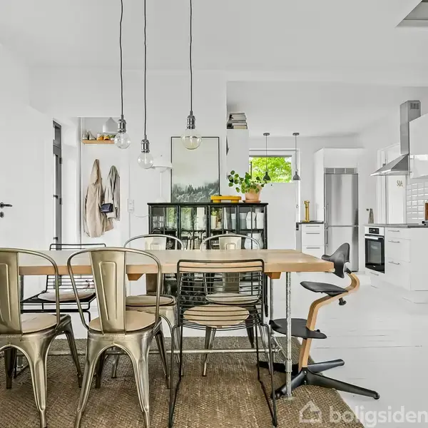 Et moderne træhus med stor terrasse i en frodig have. Havemøbler og parasoller står på en flisebelagt terrasse omgivet af græs og buske.