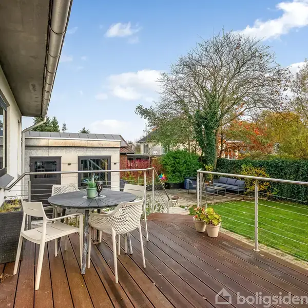 En terrasse med bord og tre stole står med udsigt over en velplejet have. Til venstre ses husets hvide facade, og baggrunden har træer og buske.