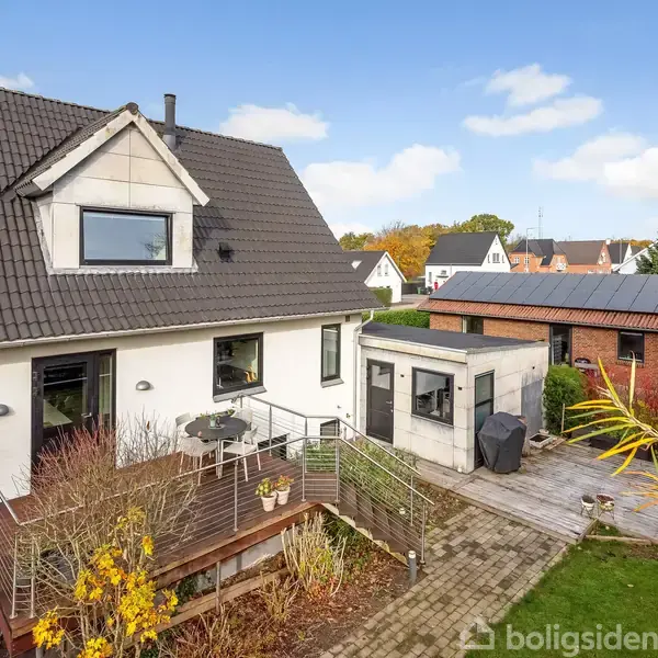 Et moderne hus med sort tag og hvid facade har en hævet terrasse med møbler. Haven er omkranset af buske. En flisebelagt sti fører til et tilstødende skur.