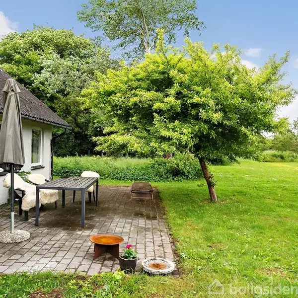 En flisebelagt terrasse ved et hvidt hus med en grå parasol og et bord med fåreskind. En grøn græsplæne omkranser området med et stort træ i nærheden.