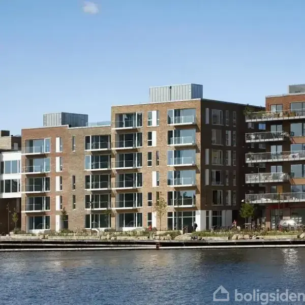 Et moderne lejlighedskompleks med store vinduespartier står langs en havnefront. En sejlbåd med et opslået sejl ses i vandet foran bygningen under en klar blå himmel.