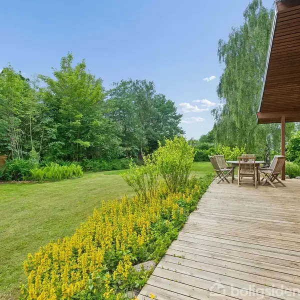 Træterrasse med bord og stole står ved siden af blomsterbede med gule blomster; i baggrunden ses en grøn have med høje træer og buske under en blå himmel.