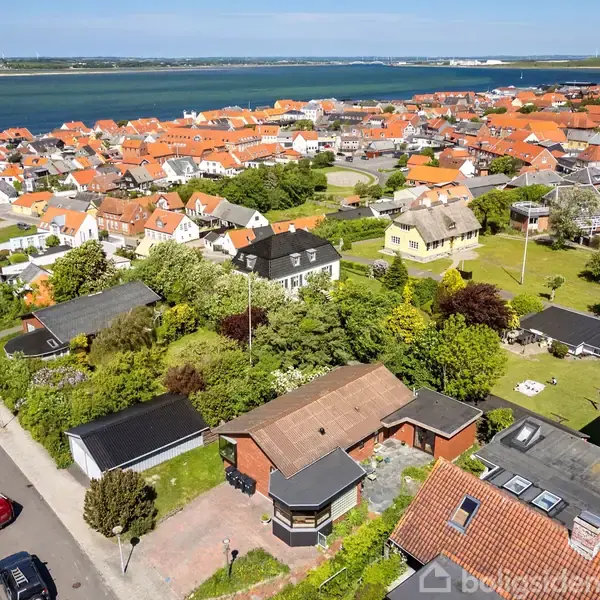 Rødt tegltagshus omgivet af grønne haver. Handling: Luftfoto af boliger. Kontekst: Kystnær by med mange huse og udsigt til vandet i baggrunden.