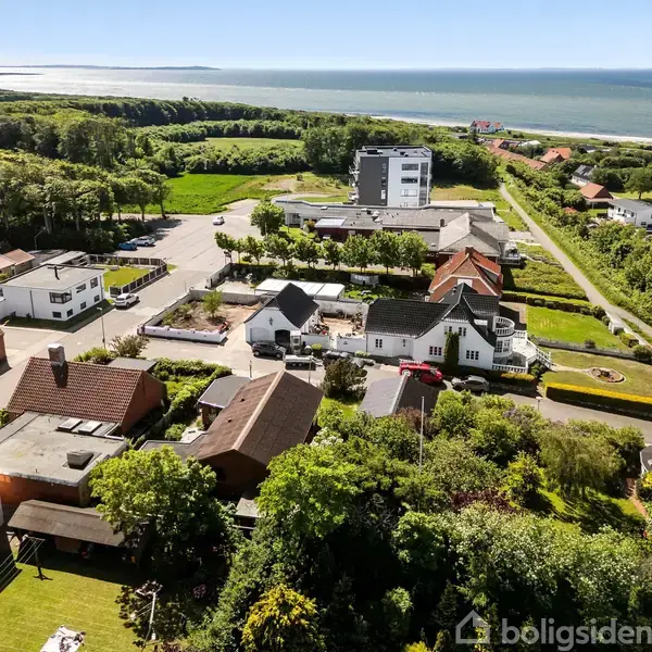 Huse med røde tage står stille i et frodigt boligområde nær kysten, omgivet af grønne træer og marker, med havet strækkende sig i horisonten.