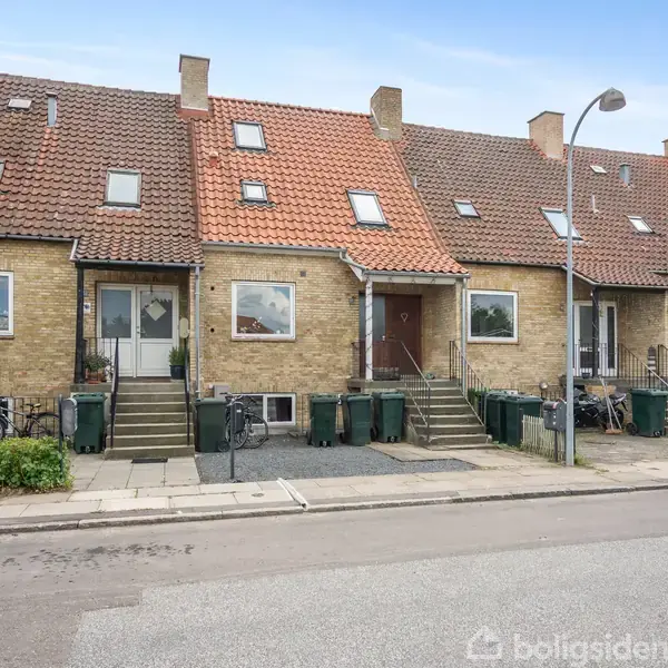 Rækkehuse med røde tegltage og brune murstensfacader har trapper ved indgangen. Skraldespande står foran husene langs fortovet. En bil er parkeret langs vejen.