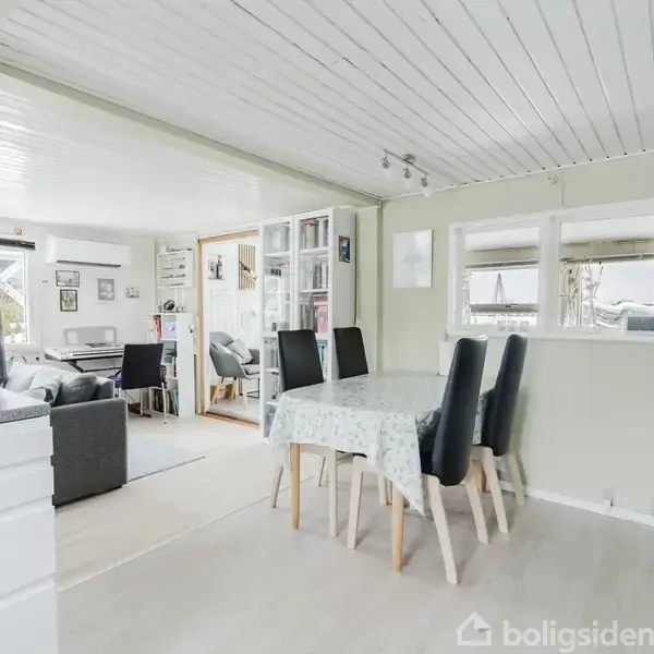 Et spisebord med fire sorte stole står i en lys stue. I baggrunden ses en grå sofa, store vinduer og dekorative billeder på væggene.