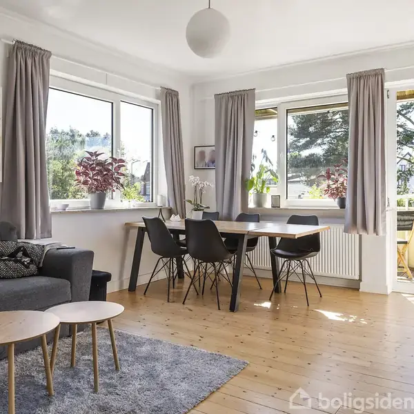 Grå sofa står i en lys stue med træbord. Spisebord til seks ved vinduerne. Gardiner hænger ved siden af vinduerne, med udsigt til have. Planter står på bord og vindueskarm.