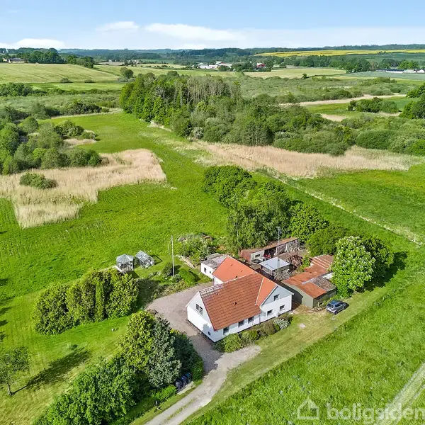 Et hvidt hus med rødt tag står i en frodig, grøn landskab. Omkring huset er der træer, marker og en grusvej, der strækker sig gennem det åbne land.