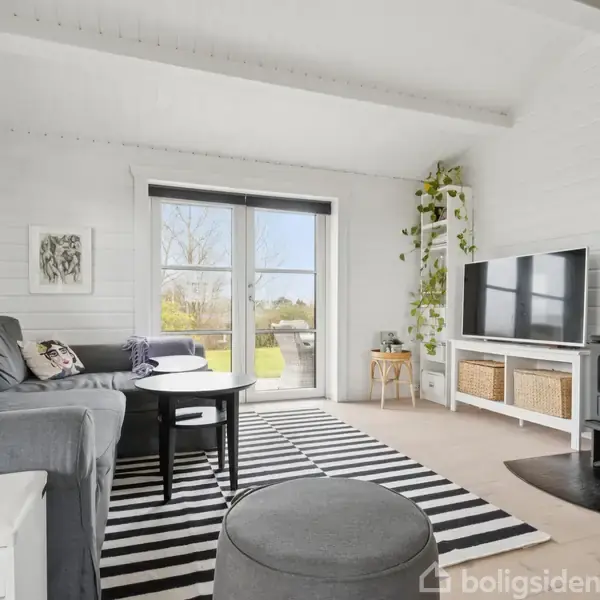Stue med grå sofa, sort pejs, og rektangulært sofabord i sorte nuancer. Stueplanter og et tæppe med striber dekorerer rummet. Store glasdøre bagest viser en have udenfor.