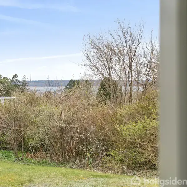 Et vindue åbner udsigt til en have med grøn græsplæne og buske, mens en træbygning ses til højre og en bakke i baggrunden under en blå himmel.
