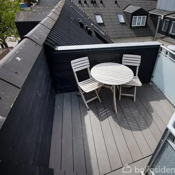 En rund hvid bordplade med to træstole står på en mørk træterrasse. Terrassen er omgivet af bygninger med mørke tage og en glasafskærmning til højre.