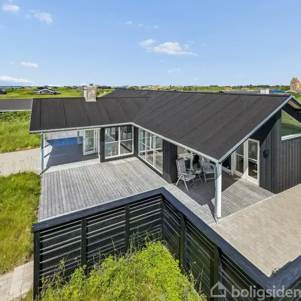 Sort træhus med terrasse, der står frit i en grøn, åben landskabsomgivelse. Terrassen har en overdækket del med stole. Den blå himmel og spredte skyer ses i baggrunden.