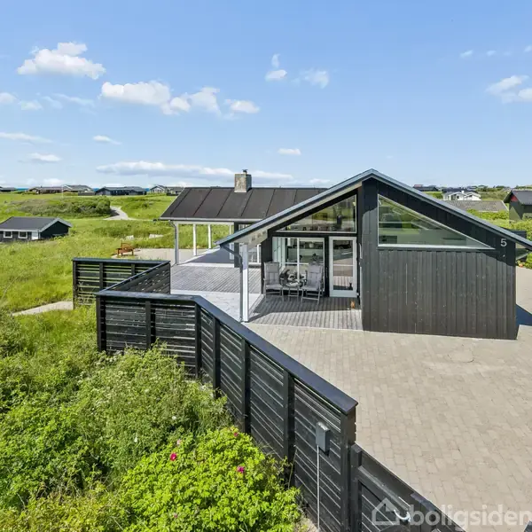 Et moderne sort træhus med store vinduer står på en flisebelagt terrasse. Huset er omgivet af grønne marker og andre spredt beliggende huse under en blå himmel med skyer.
