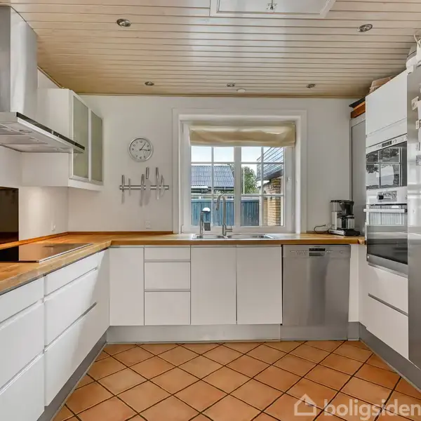 Køkkenet har hvide skabe og træbordplade, med en opvaskemaskine og køleskab til højre. Et ur og knager hænger på væggen, og et vindue giver udsigt til en terrasse.