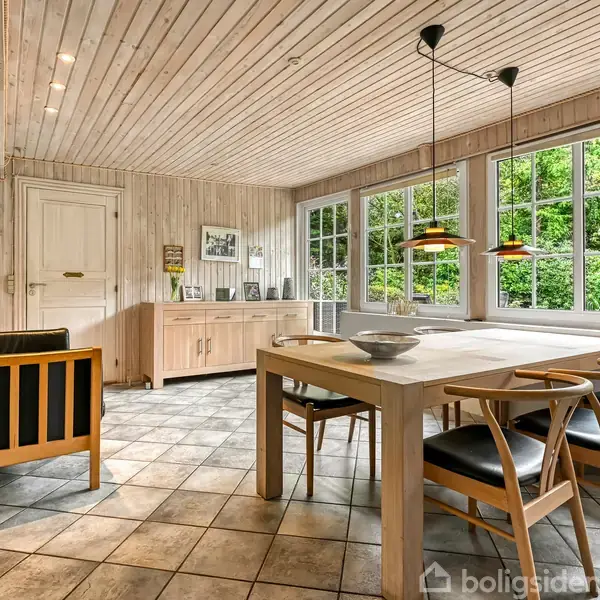 Spisestue med træbord og stole, sofahjørne med sort sofa til venstre, lyset flyder ind fra store vinduer. Et hvidt skab står ved bagvæggen med billeder og dekoration.