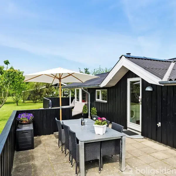 Terrassebord med stole står under en stor parasol. Bordet har blomsterdekorationer. Den sorte træbygning ligger i et grønt haveområde med træer omkring.