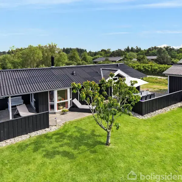 Et sortmalet fritidshus står på en velplejet grøn græsplæne. Huset har en stor terrasse omgivet af et lavt hegn. Omgivelserne er landlige med andre huse i baggrunden.