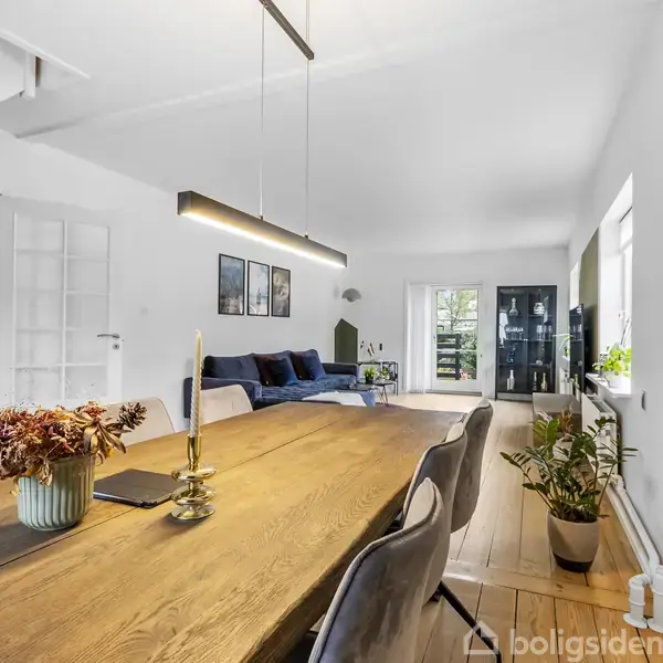 Langt træbord med stole arrangeret omkring det i en moderne spisestue. Et hængende lys over bordet, med en åben stue i baggrunden, der indeholder sofaer og planter.