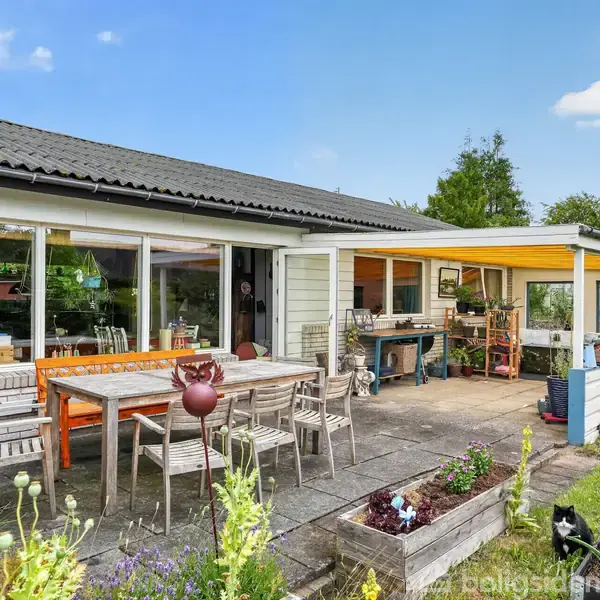 Hus med én etage har en udendørs terrasse. Der står et træbord med stole på terrassen, som er omgivet af planter og blomsterbede i en grøn have.