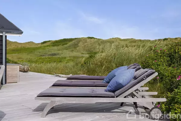 Liggestole med hynder og blå puder står på en træterrasse. De er placeret ved siden af et hus. I baggrunden er der grønne klitter og en blå himmel.