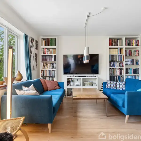To blå sofaer står overfor hinanden i en stue med trægulv. Et tv er monteret på væggen mellem bogreoler, og et sofabord står centralt. Store vinduer lader lys strømme ind.