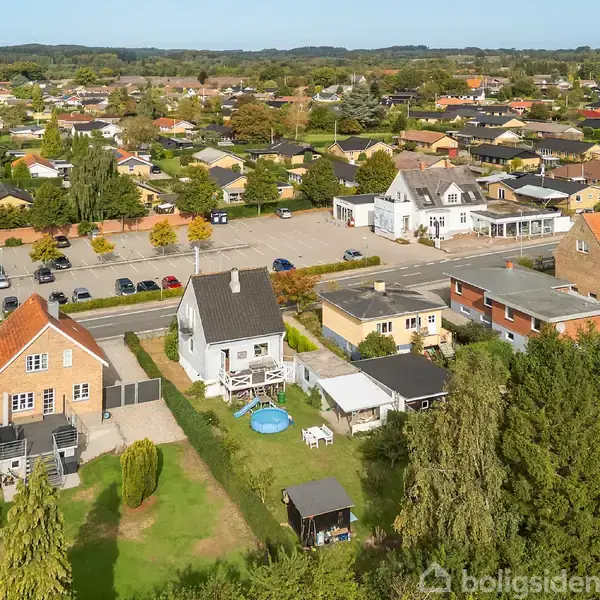 Et parcelhuskvarter med huse og haver set fra oven. Et hvidt hus med pool i haven står i forgrunden. Omkringliggende bygninger og træer danner en forstæderne atmosfære.
