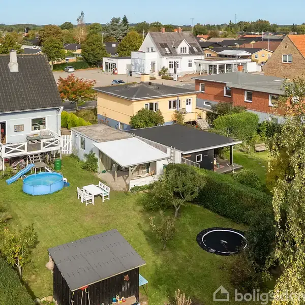 Et parcelhus med en have indeholder en blå oppustelig pool og en trampolin. Der er et haveskur i forgrunden. Omgivelserne viser flere boliger og træer i et parcelhuskvarter.