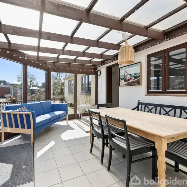 En lys udestue med et træbord omkranset af sorte stole og en blå sofa. Skydedøre i glas fører ud mod en have med udsigt til et rødt hus.