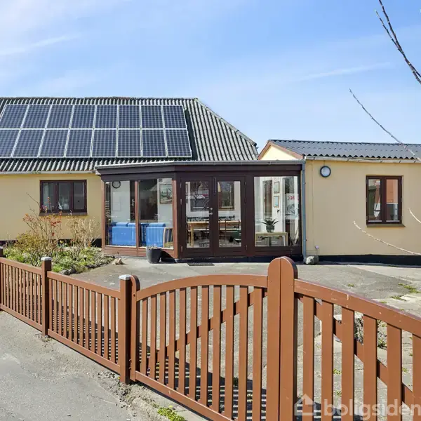 Et hus med solpaneler på taget. Foran huset er der en indkørsel og en træport i et villakvarter. Der vokser et træ ved siden af indkørslen.