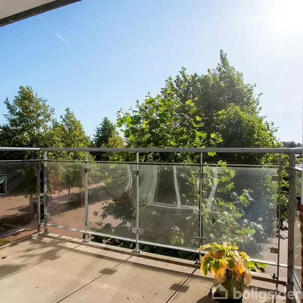 Balkon med gennemsigtigt gelænder. En potteplante står tæt på kanten. I baggrunden ses frodige grønne træer under en klar, blå himmel. Solen skinner fra højre.