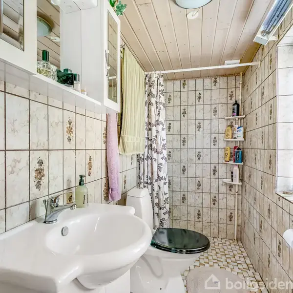Vask med hvid porcelæn står under et spejlskab. Fliser med blomsterbeklædning omgiver bruser og toilet. Vinduet til højre lader lys ind i det træloftede badeværelse.