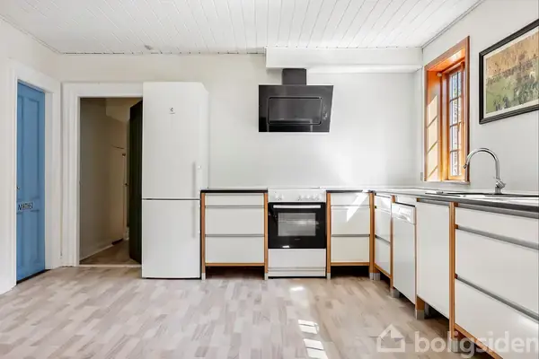 Et køkken med hvide skabe og trækanter, et komfur under en sort emhætte, køleskab til venstre, opvaskemaskine på højre side; dagslys skinner gennem et vindue.