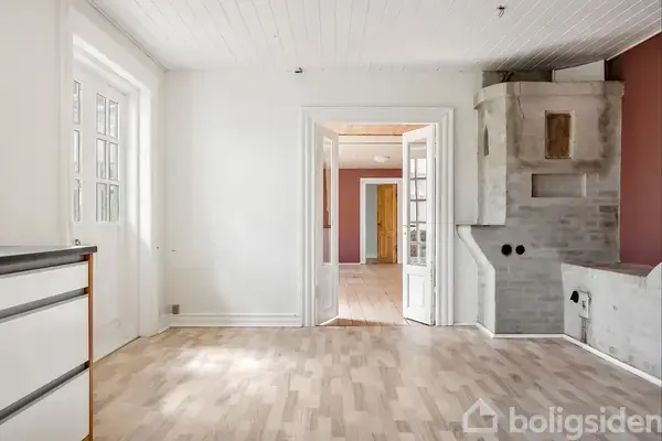 Et lyst rum med trægulv. Dobbeltdøre åbner ind til et tilstødende rum. Der er en grå brændeovn til højre op ad væggen.