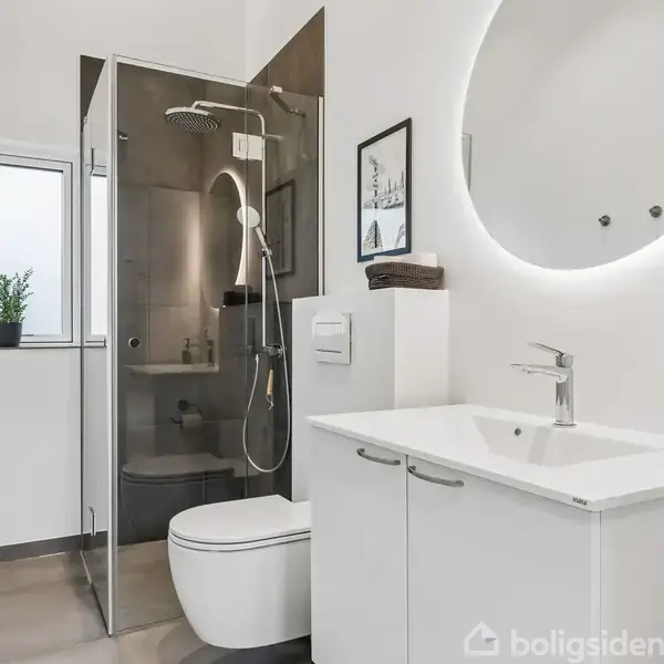 Badeværelse med hvidt skab og håndvask, bruser i glasaflukke. Rundt spejl hænger over vasken. Plante ved vindue, vægudsmykning over toilettet. Rummelig og moderne indretning.