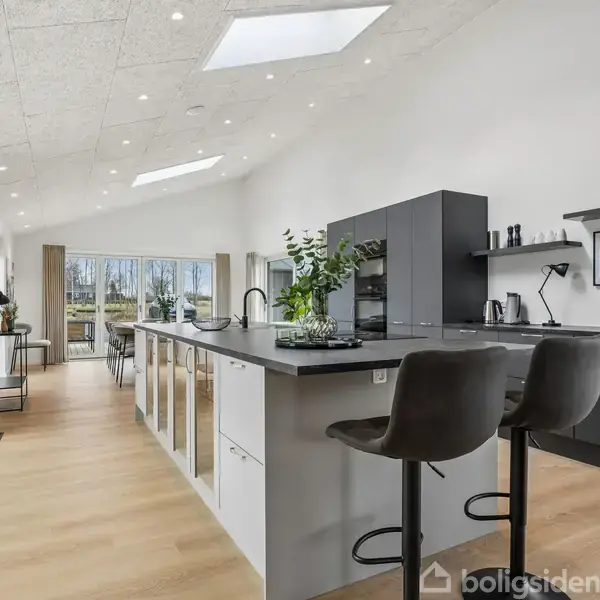 Et moderne køkken med en stor ø midt i rummet. Der står to barstole ved øen, og elegante grå skabe er langs væggen. Store vinduer til venstre giver naturligt lys.