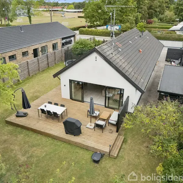 Et hvidt hus med skråt tag har en stor træterrasse med havemøbler og en grill. Haven omgiver huset med græs og træer, og nabohuse ses i baggrunden.