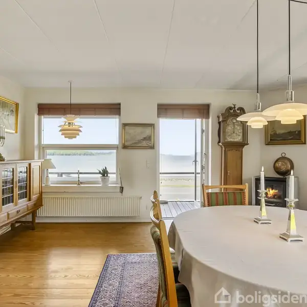 Spisebord med lysekrone over, omgivet af stole i en lys stue. I baggrunden ses et skab, billeder på væggen og en udsigt til stranden gennem vinduerne. Ingen tekst.