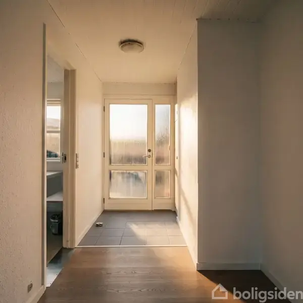 En lys gang fører mod en glasdør, der lukker sollys ind. Til venstre er der et baderum. Væggene er hvide, og gulvet skifter fra træ til fliser.
