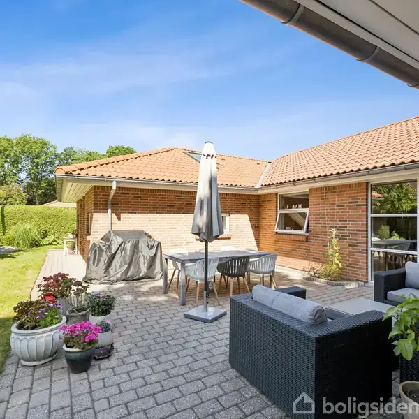 En udendørs terrasse med havemøbler og parasol. Stenklinker dækker jorden. Grønne planter og buske omgiver området, ved siden af et murstenshus med rødt tegltag under en blå himmel.