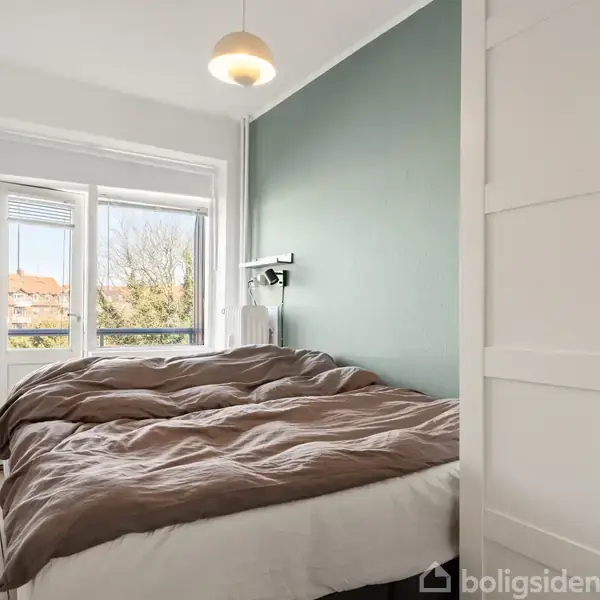 Soveværelse med dobbeltseng dækket af brunt sengetøj, placeret ved grøn væg ved siden af skab. En lampe hænger over sengen, og et vindue viser udsigt til træer og bygninger.