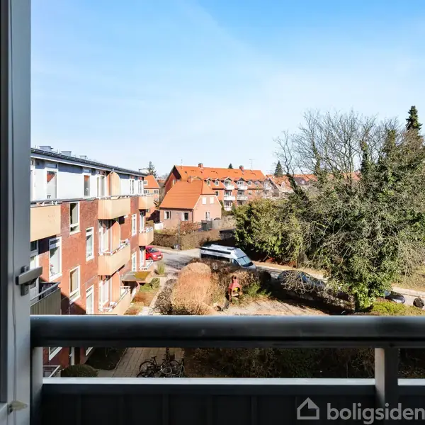 En altan viser udsigt over en gård mod røde bygninger og et træ. Flere biler er parkeret langs vejen. Klar blå himmel skaber en lys atmosfære.