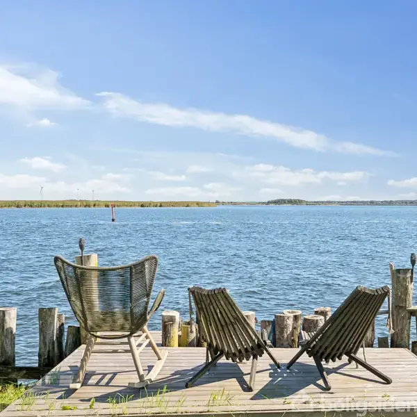 To stole står stille på en træterrasse med udsigt til en rolig sø omkranset af træstolper, under en klar blå himmel.