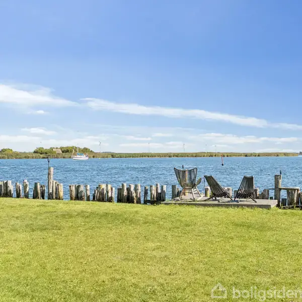 Grønt græsplæneområde, der grænser op til træpæle ved vandkanten, hvor to liggestole og et bålsted er placeret under en blå himmel.