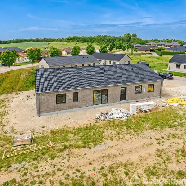 En enetages murstenshus med mørkt tag står på en åben byggeplads, omgivet af græs og grus. Omkring huset ses flere huse, samt en bil på en grusvej.