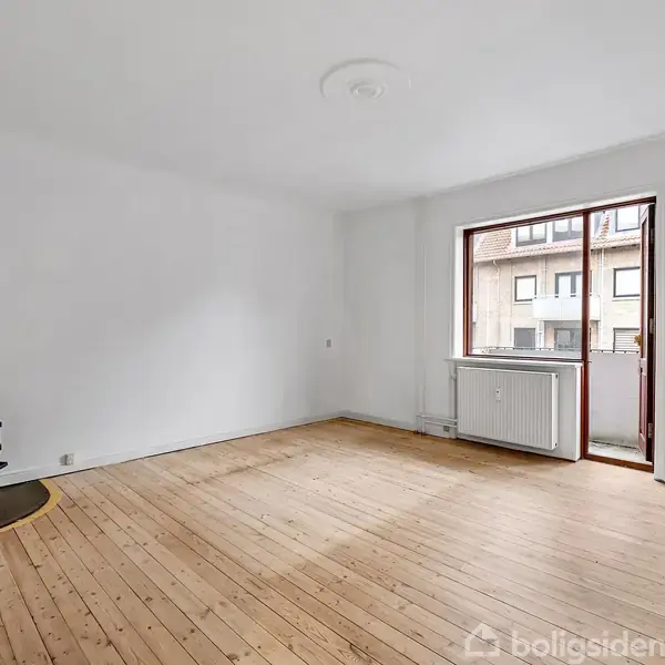 Et tomt rum med trægulv, en sort brændeovn placeret i hjørnet. Store vinduer og en dør til altanen giver lysindfald. Væggene er hvide uden dekorationer.